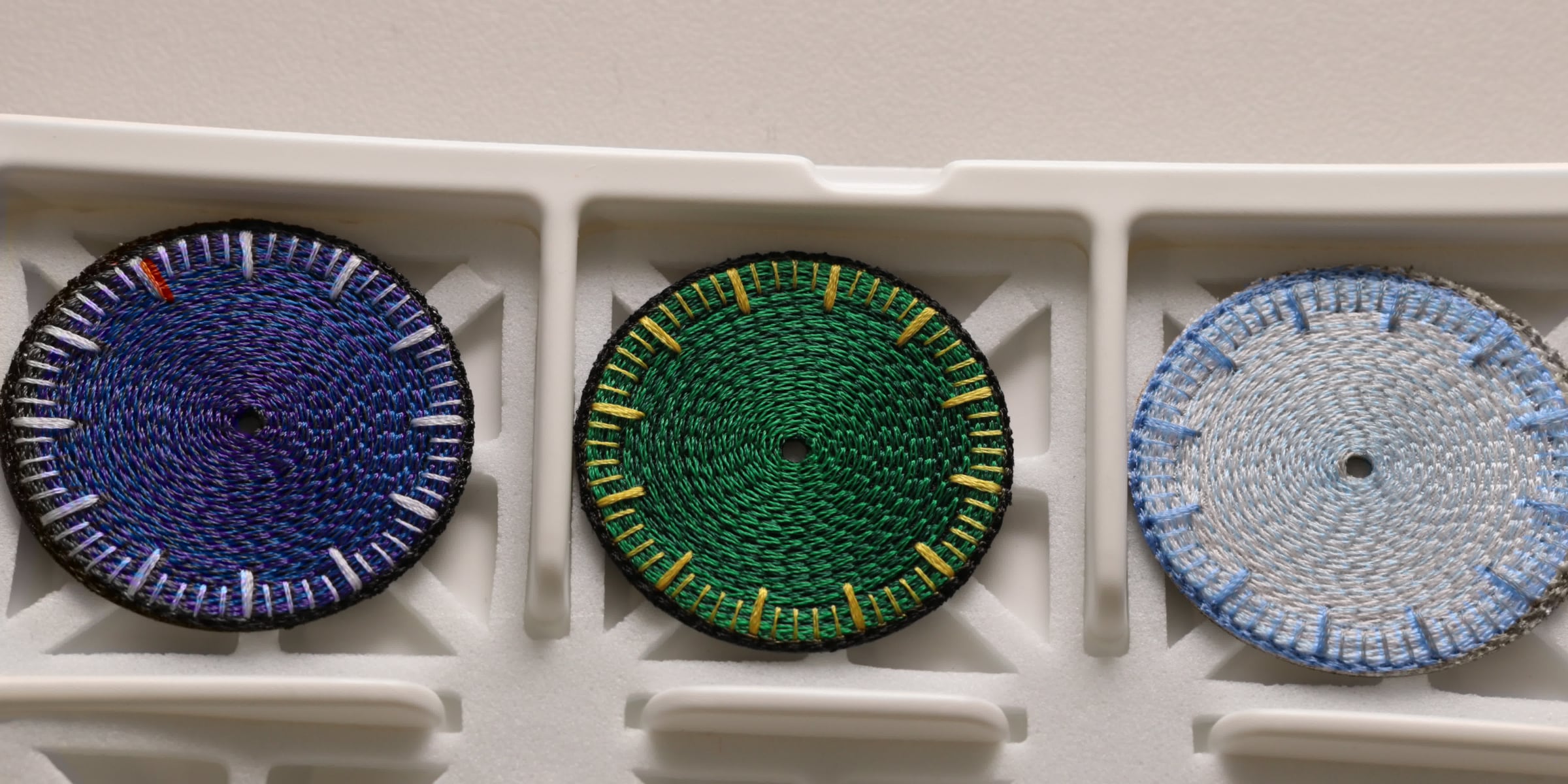 Three chenille-embroidered watch dials, deep blue-violet, green and pale blue, resting in a white production tray