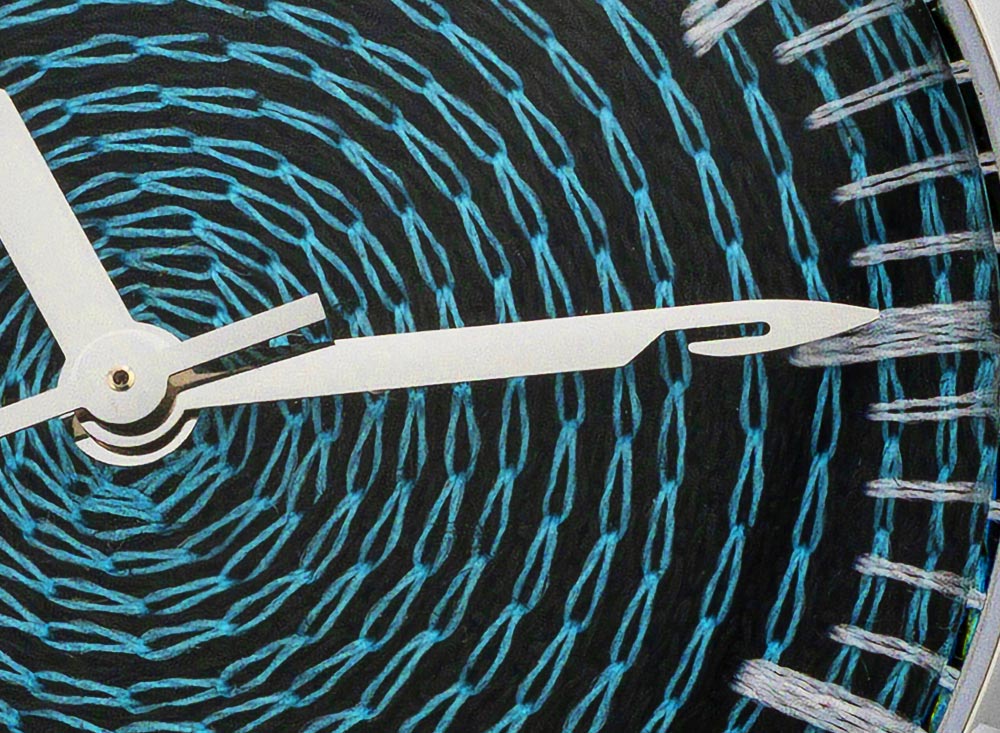 Extreme macro of chenille-embroidered watch dial, teal thread spiral and needle-shaped hands